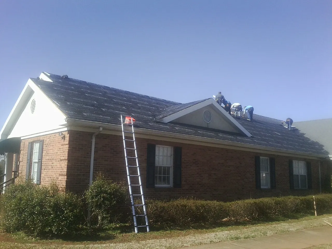 Crew tearing shingles off a brick ranch roof with a ladder set — Metal Roof Repair work by our crew in Chatham